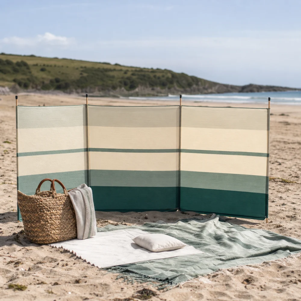 A beach windbreak set up beside towels and a beach bag on sand.